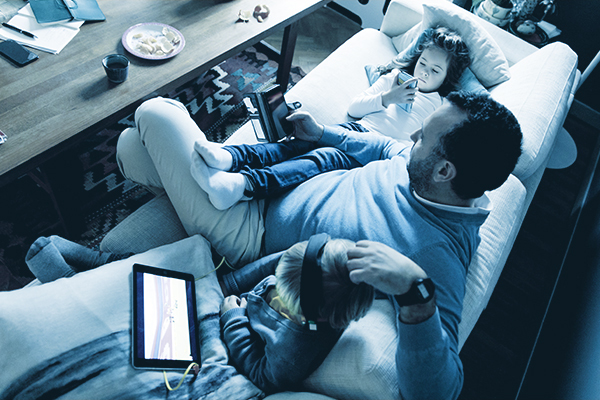 600x400_DuotoneImages_HomeProtector2 Dad seating on the couch with his kids, all three playing on electronic devices